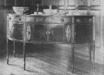 This fine Sheraton sideboard shows curved doors, and
knife boxes with oval inlay of satinwood. The center cupboard is
straight. The legs are reeded.